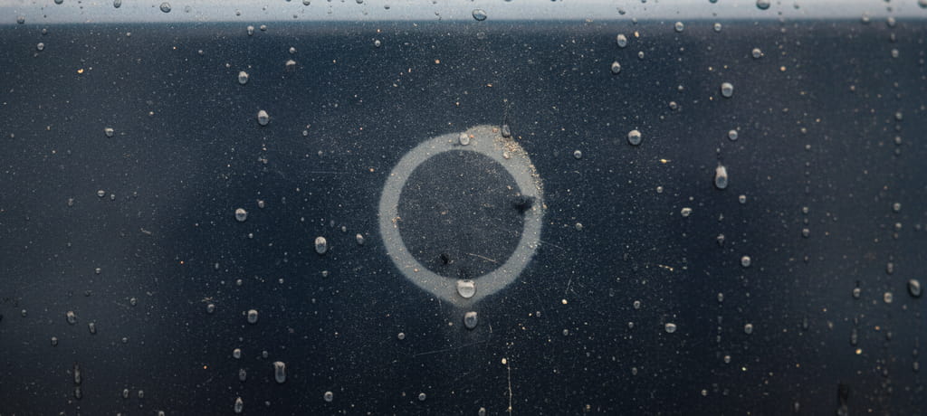 Close-up view of a car door surface with raindrops and dust particles catching the light, a faint circular outline where something once rested, symbolizing wear, time, and the delicate balance between protection and exposure, moody natural lighting, no text
